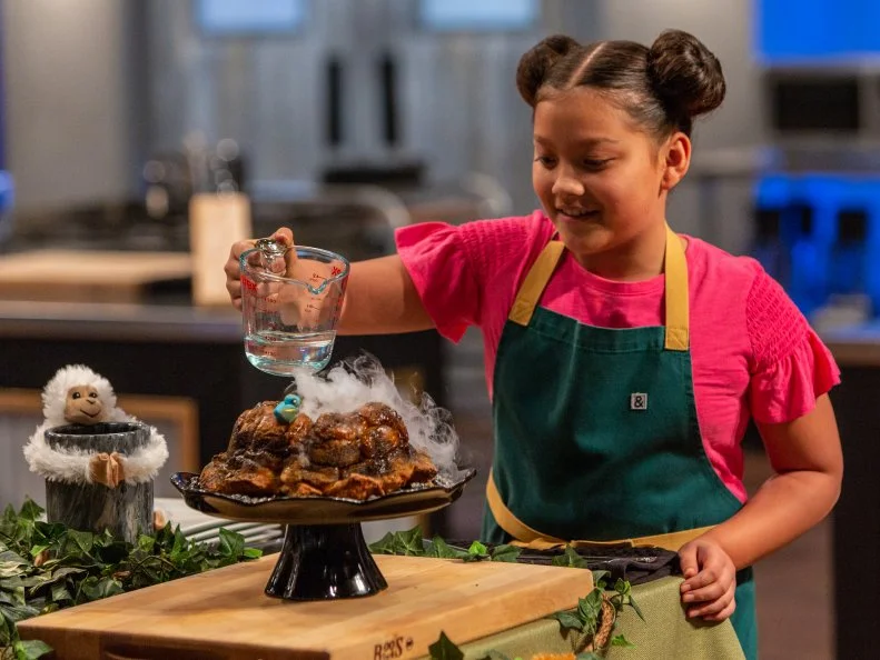 Contestant Meadow Roberts pours water onto her dry ice in her dish for a volcano effect, as seen on Kids Baking Championship, Season 6.