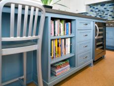 A large, blue kitchen island not only offers room for prep and cooking but also features seating, built-in cookbook storage, drawers and a wine refrigerator. A metal chair has a modern look against the contemporary cork flooring and blue tile backsplash. 