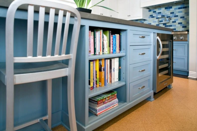 A large, blue kitchen island not only offers room for prep and cooking but also features seating, built-in cookbook storage, drawers and a wine refrigerator. A metal chair has a modern look against the contemporary cork flooring and blue tile backsplash. 
