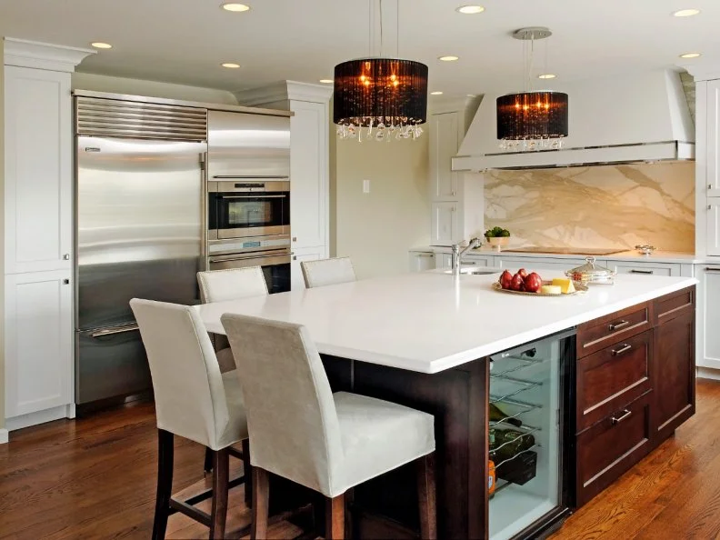 In this contemporary kitchen, two chandeliers balance the length of the large island and raise the eye upward. The marbled backsplash complements the tan wall and brown island cabinets. Large islands are the ideal space to add much-needed storage as well as a few luxuries. Designer Jennifer Gilmer of Jennifer Gilmer Kitchens & Bath Ltd., included an under-counter beverage refrigerator, deep and shallow drawers, and even an extra prep sink to create a functional and stylish island. 