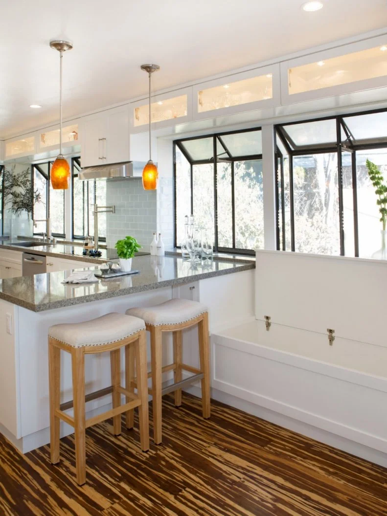 Gorgeous angled windows fill this bright and airy kitchen with lots of natural light. A pair of upholstered wood barstools provide an eat-in spot at the countertop, and a nearby window seat offers additional seating (or storage). Bold orange pendant lights and beautiful striped hardwood floors add finishing touches to the design.