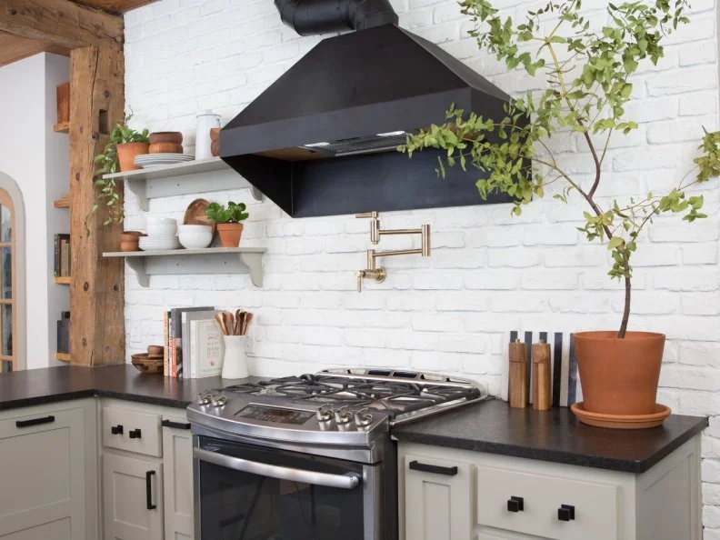 The home's original shiplap is exposed on the kitchen ceiling. A brick paver backsplash painted in white provides a contrasting backdrop for the black vent hood, dark granite countertops and distressed tile flooring.
