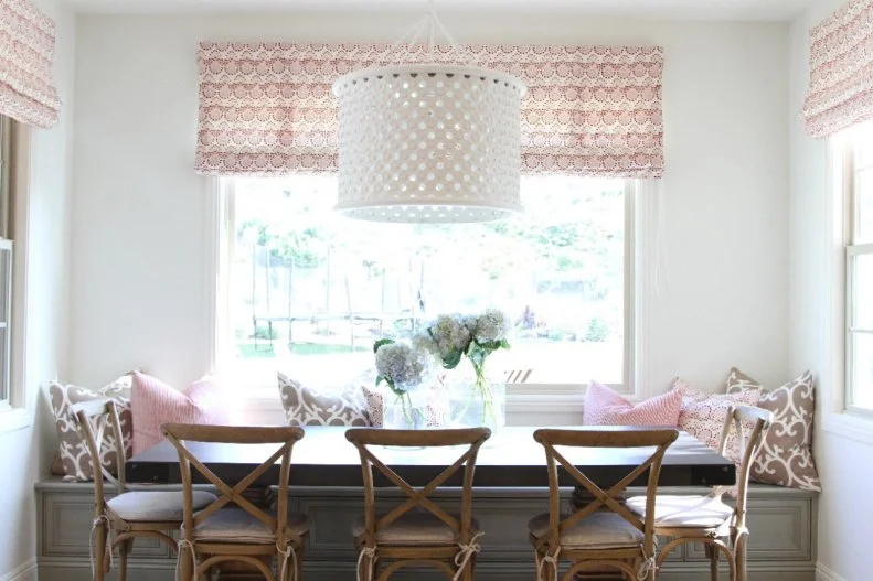 The coastal breakfast nook is framed by windows topped with pink block print window treatments. Charming banquette seating is paired with a rustic table and chairs, and an airy white drum pendant is an eye-catching focal point. The finished result is an inviting, clean and colorful nook. 