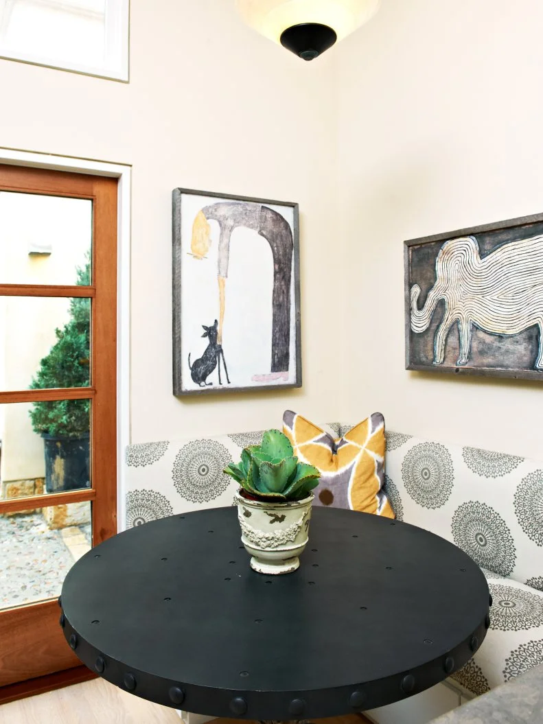 A black and white upholstered banquette sets the tone in this eclectic breakfast nook complete with a round dining table, framed artwork and natural lighting. 