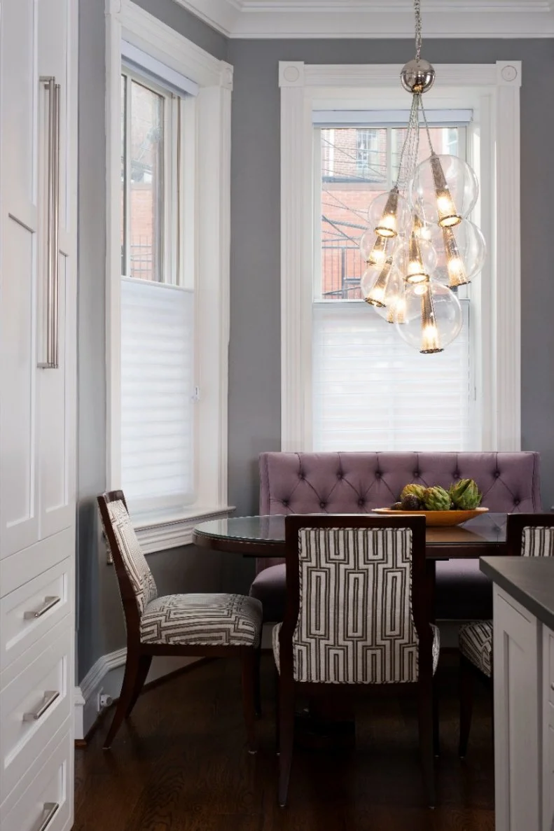 A cluster of globe pendant lights hangs above the breakfast nook table, adding a fresh, modern look to the transitional space. The tufted lavender purple settee injects a sophisticated pop of color, while graphic patterned chairs bring a playful touch to this dining area.