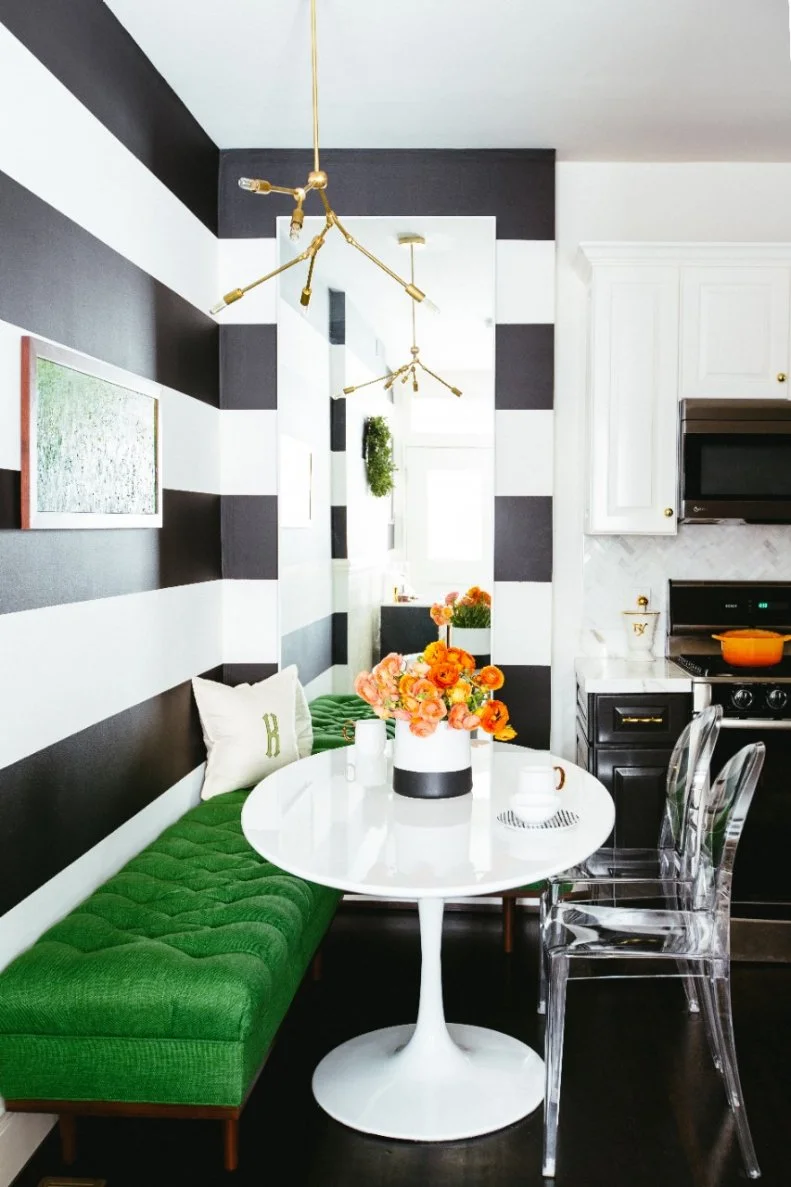 Painting the walls in a high contrast black and white stripe tricks the eye into seeing more light than the space actually has. The black and white motif is repeated in the kitchen cabinets with added subtle touches of brass via the light fixture and cabinet hardware. A corner L-shaped bench adds a pop vibrant kelly green that works perfectly with the wall art piece.
