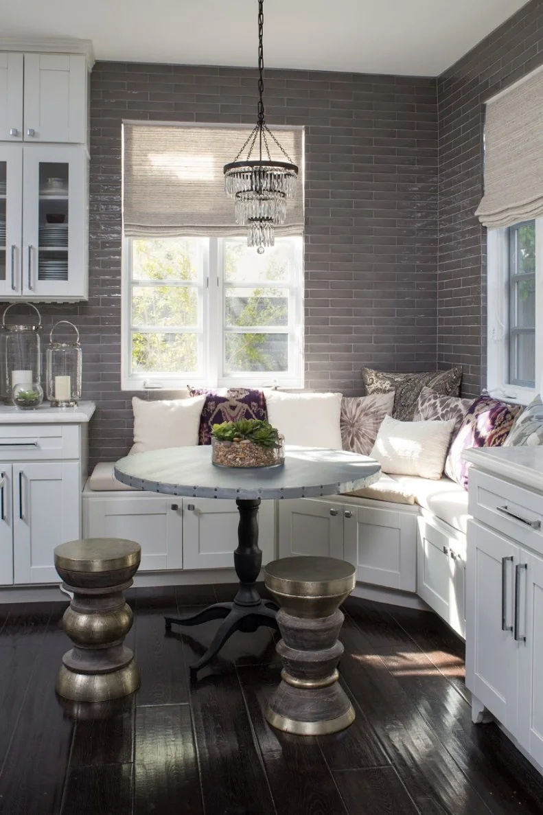 A custom corner banquette and round pedestal table make for a cozy breakfast nook in this gray-tiled eat-in transitional kitchen. Cabinets below provide handy built-in storage, while a pair of stools offers extra seating.