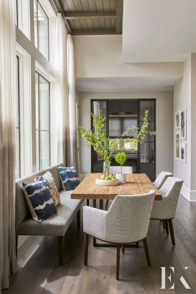 The cozy breakfast nook pairs a rustic dining table with a sleek bench and comfortable armchairs. Exposed wood beams reflect the hardwood floors of the space. 