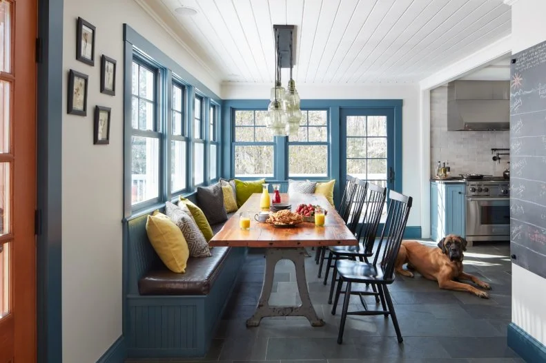 To define the breakfast nook, the banquette and surrounding trimwork is painted a striking blue. Classic Windsor chairs sit opposite the leather-cushioned banquette, surrounding the farmhouse-inspired table. 