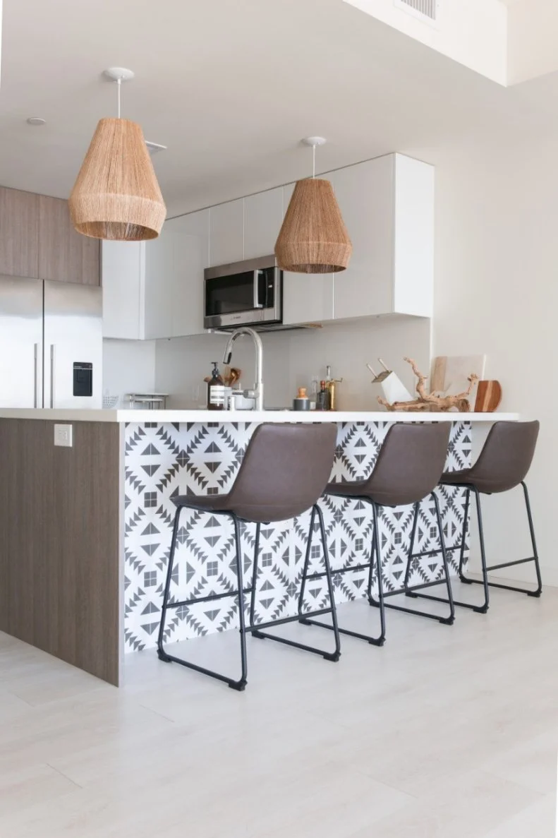 Grass-woven pendant lights and driftwood accent add a natural, beachy feel to the breakfast bar in this kitchen. The light tones blend well with the medium tones of the cabinetry and dark brown bar stools.