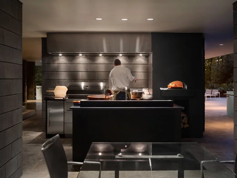 An outdoor kitchen area includes a grill, wood-fired oven and island for prep at this modern Dallas home. The kitchen is centrally located in the home's public entertaining area near the pool deck and open-air dining area.