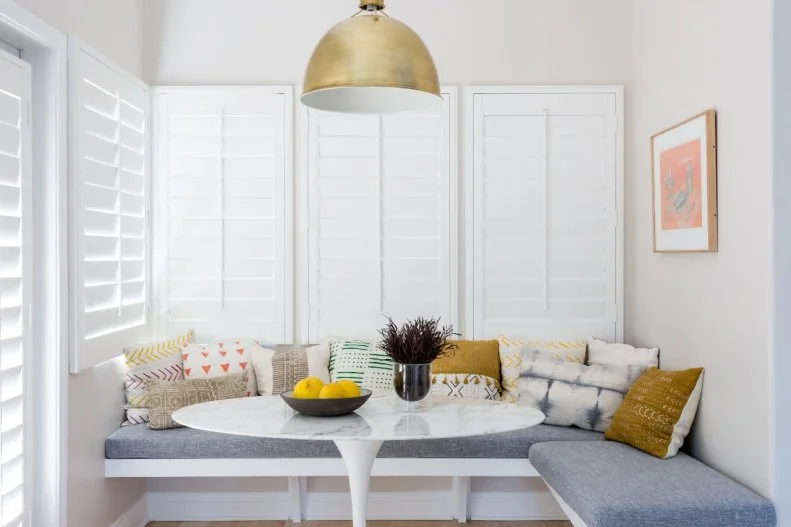This modern white breakfast nook features a gray upholstered built in banquette with colorful textile accent pillows, a midcentury modern marble topped tulip table, and gold pendant light.