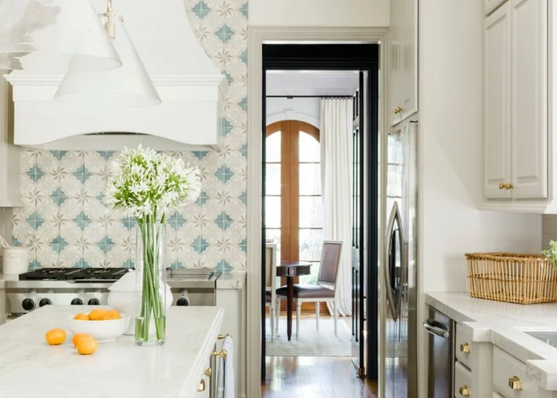 This traditional kitchen is elegantly decorated with white cabinets with gold hardware, white range hood, marble countertops, and a soft blue and gray mosaic tile backsplash.