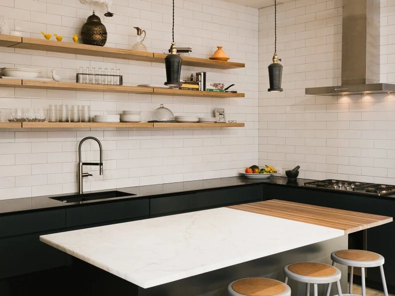 The modern kitchen features wall-to-wall oversized subway tiles paired with black cabinets and countertops for an eye-catching contrast. Wooden wall shelving above the sink provides room for storing kitchen essentials, while a white marble kitchen island with a partial wood section allows extra room for food preparation.