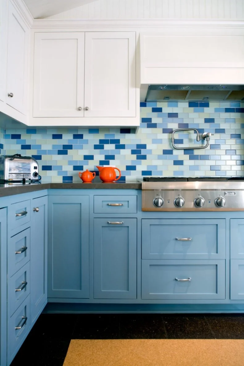 A backsplash of blue and pale yellow subway tile complements the dark gray countertops while bringing in pops of color. A mix of white and blue cabinets add contrast to the space, while a cork floor provides a unique textural element in this transitional kitchen. 