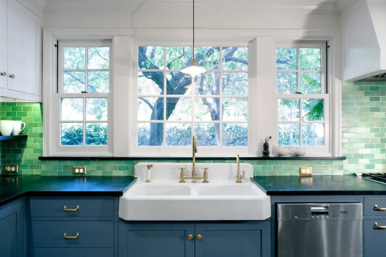 Green subway tiles adds a playful pop of color to this transitional kitchen, contrasting nicely with the white farmhouse sink and cabinets. A large window and pendant light ensure that the room is airy and bright, no matter what time of day it is. 