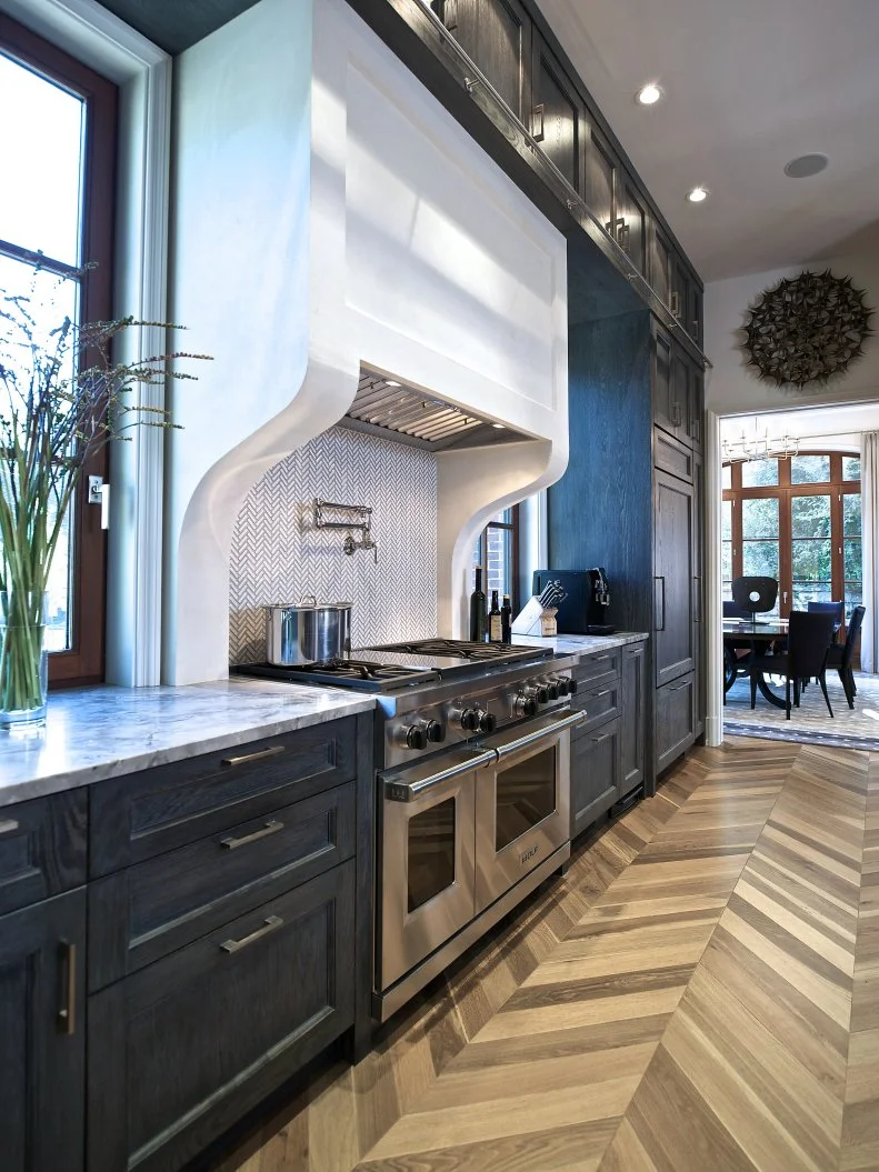 A crisp white range hood pops against the dark bluish black cabinets in this transitional kitchen. Chevron patterned wood floors and a coordinating ceramic tile backsplash create movement and interest within the space.