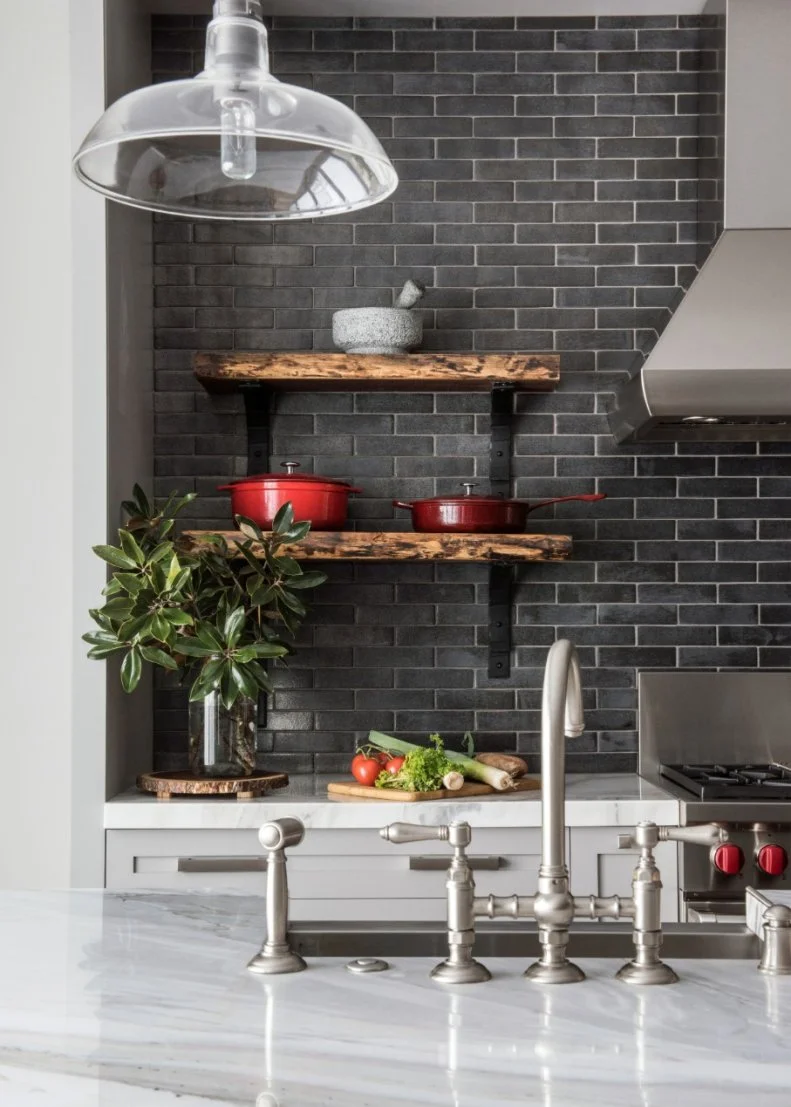 This stylish kitchen features a gray subway tile backsplash paired with white and gray marble countertops. Brushed nickel fixtures line the kitchen island sink, while a glass pendant light hangs above. Built-in wooden shelves provide the perfect spot for displaying bold red cookware.