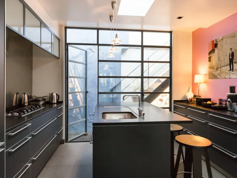 A splash of pink adds interest to the otherwise monochromatic kitchen, featuring sleek cabinets and a center island with a sink. Glass doors open to an outdoor staircase, which leads to a spacious rooftop terrace.