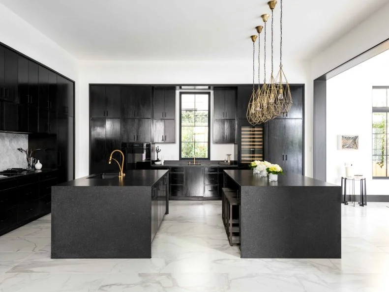 Black cabinets, island and dining table bring dramatic flair to the bright white walls and marble floors in this open floor plan kitchen. Gold pendant lights bring a touch of metallic over the sleek, built-in dining table, and the entire space opens up to the living room, making it perfect for entertaining. 