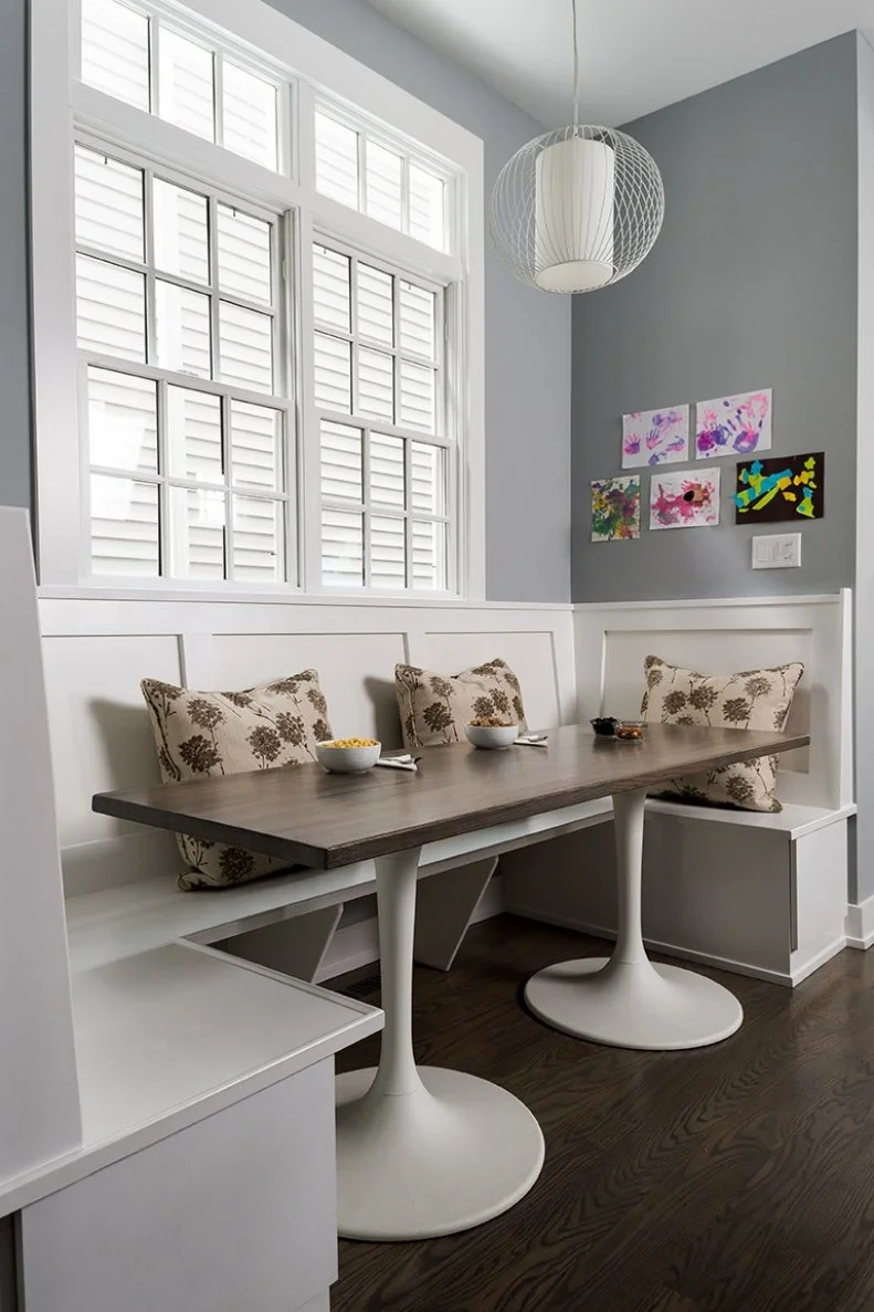 This contemporary breakfast nook features a custom midcentury-modern-style dining table and bright white banquette. Pillows in brown tones bring a soft, natural feel to the space, and a round light fixture echoes the circular table bases, pulling the room together.