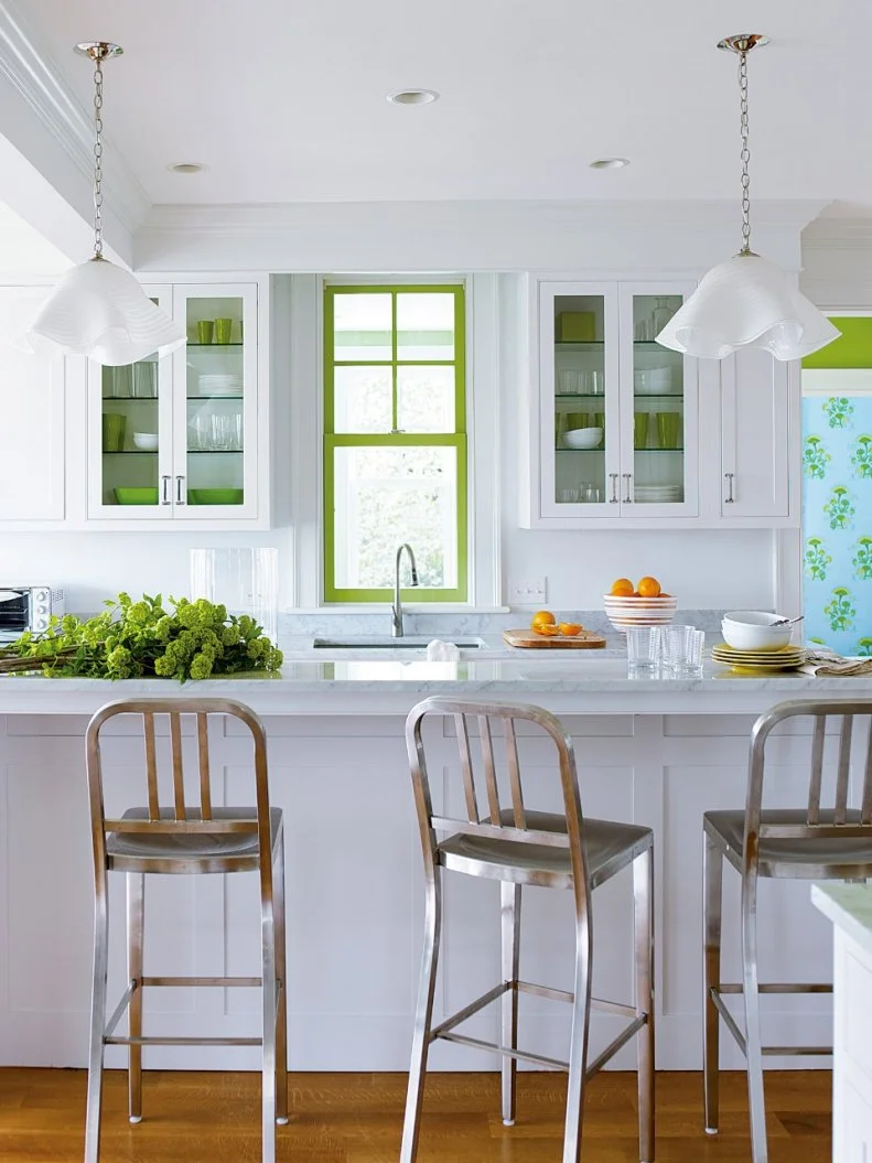 Green accents pop in this white, cottage style kitchen featuring floral wallpaper. Metallic stools at a breakfast bar add a contemporary touch. 