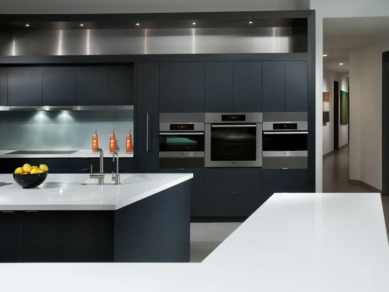 Designer Angelica Henry used a low maintenance approach and a sleek, stylish aesthetic when designing this black, white and stainless steel kitchen. Miele appliances, linear overhead lighting, black laminate cabinets, white Silestone countertops, a glass backsplash and a stainless steel soffit all help to define its intended architectural character.
