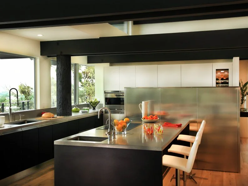 The contemporary kitchen features a black-and-white color palette with stainless steel accents. The large black island with a stainless steel countertop provides bar dining with sleek barstools. The original 1970s house was constructed on a series of telephone poles cantilevered off the canyon floor, so they become a unique part of the interior design.