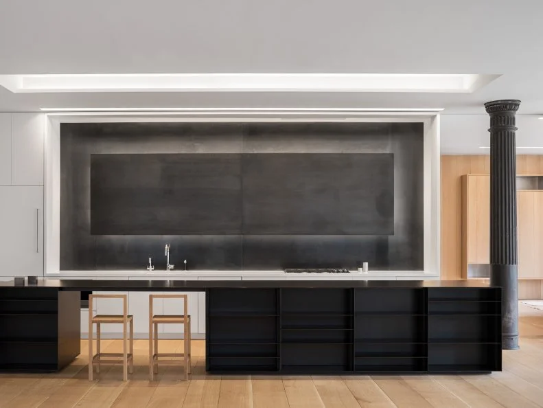 Raw steel is used for the backsplash and island in a loft's open kitchen. The backsplash is lit by LED lights for a halo-like effect, and the island has shelving on both sides in the black-and-white kitchen. The project by Desai Chia Architecture won a 2018 AIA Institute Honor Award for interior architecture.