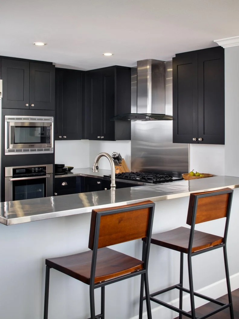 Black cabinets create a bold contrast to the extremely pale gray walls and white walls and ceiling in this kitchen space. Stainless steel appliances are further tied into the design with stainless steel countertop. Masculine, modern wood bar chairs add a touch of warm to the cool tones of the room.