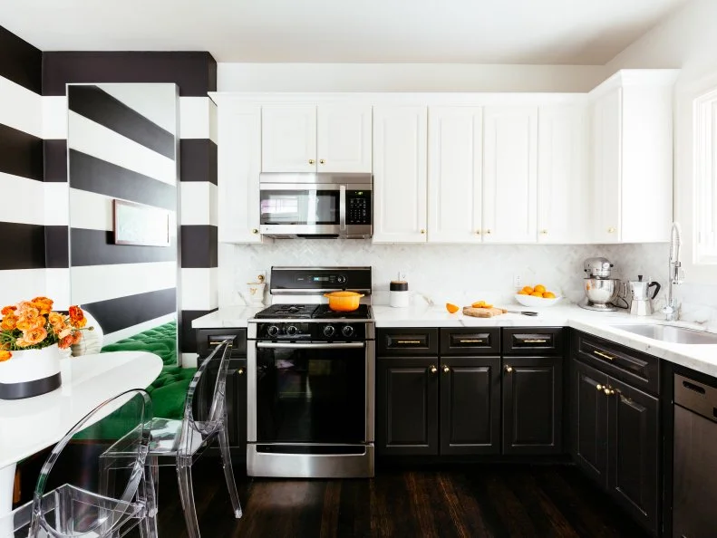 A marble herringbone backsplash separates the white marble countertops and built in white cabinetry. Lower black cabinets create a beautiful contrast to the white and echoes the thick black and white wall stripes. The black and white creates a light and dramatic look. Clear plastic chairs sit at the modern white dining table decorated with a bright orange bouquet. 
