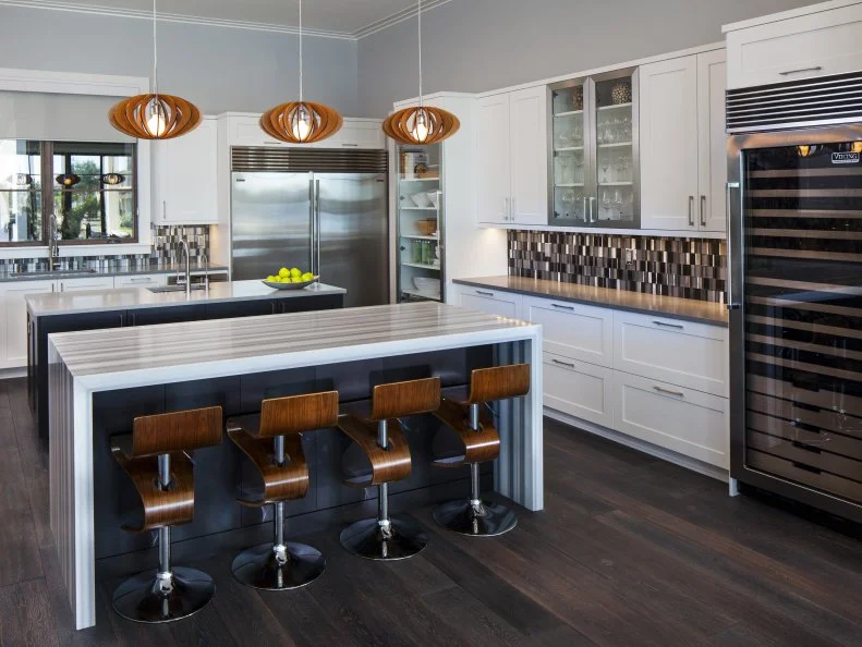Working with a monochromatic color palette of gray, black and white, the kitchen boasts bright white cabinets, two kitchen islands in black and dark wood flooring. Four, dark wood barstools line the island with a waterfall granite countertop to offer a place to dine. A large wine fridge is a great addition for the host who loves to entertain.