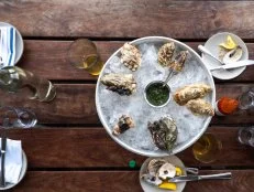 A plate of oyster shells overturned on a bed of ice sitting atop wooden table. The beer, hot sauce, and lemon wedges that complimented the meal scatter the table.