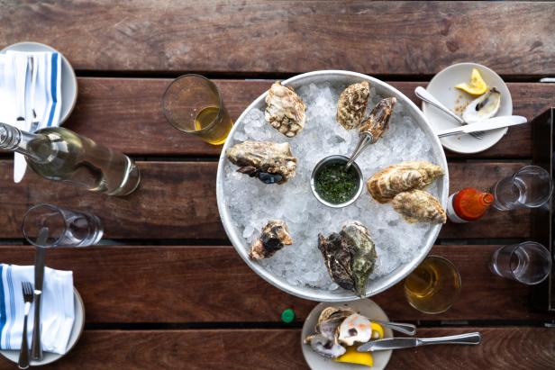 A plate of oyster shells overturned on a bed of ice sitting atop wooden table. The beer, hot sauce, and lemon wedges that complimented the meal scatter the table.