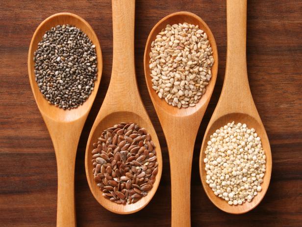Four light-colored wooden spoons filled with different types of seeds positioned on a dark wood table. From left to right, the spoons contain chia, flax, sesame and quinoa seeds.  They are arranged with the handle facing in the opposite direction with every other spoon.
