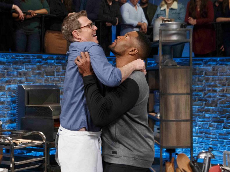 Co-host Michael Strahan lifts host Bobby Flay as he cooks Chicken and Waffles during round two, as seen on Beat Bobby Flay, Season 19.