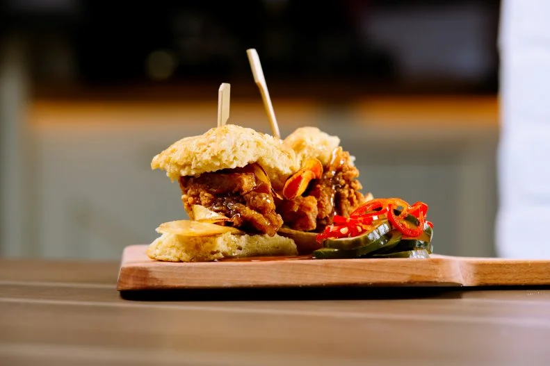 At their Miami and New York City restaurants, husband-and-wife Top Chefs Jeff McInnis and Janine Booth serve a fun and delicious fried chicken dish with layer upon layer of bold flavors and textures. Their crispy fried chicken is brined in sweet tea, battered, tossed in zested lemon powder, drizzled in pepper jelly and wrapped in a buttery and flaky angle biscuit. One would think that would be enough for most chefs, but these two decided to take it up another couple of notches. “I love adding potato chips to sandwiches,” says Booth, “so we decided to add our housemade root chips for extra crunch and our homemade mix of pickled cucumbers, red onions and Fresnos for acidity and sweetness.”