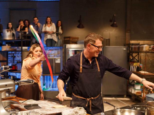 Co-host Damaris Phillips waves a ribbon at chef Bobby Flay while he tries to prepare his Round 2 dish, as seen on Beat Bobby Flay, Season 24.