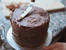 A close-up photograph of a girl decorating a cake with chocolate ganache in a domestic kitchen