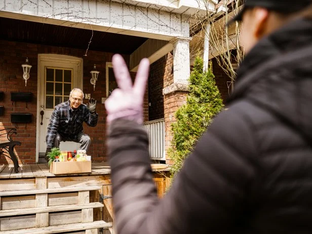 COVID-19, Young women delivers fresh food to senior man during pandemic lock-down. Senior man picking up the box of fresh produce off the porch while woman is greeting with the peace sign from the distance. Urban setting of a North American city.