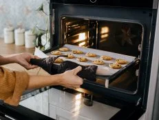 Young mother is putting a tray full of cookies which they made together with her husband and her son in the oven. Horizontal photo. Face is not visible.