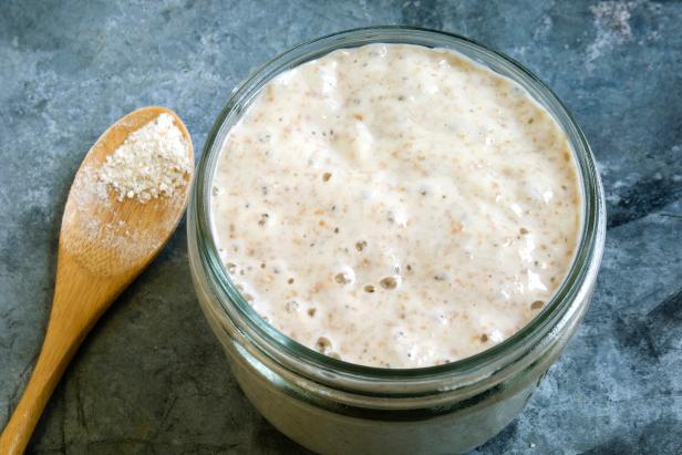 A closeup of sourdough starter with wooden spoon and wholewheat flour on metal table top.