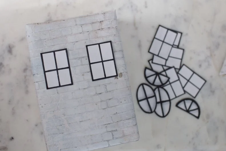 Now it’s time to add the decorative accessories. Draw windows and doors on colored and white paper. You can do this freehand or you can print out windows and outline them with your black marker. Cut them out and set aside. Cut a few strips from your scraps of paper to make the shutters too. 