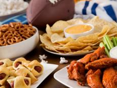 Tailgate party spread with hot wings, pig in a blanket, nachos, pretzels, and popcorn with football and a football jersey.