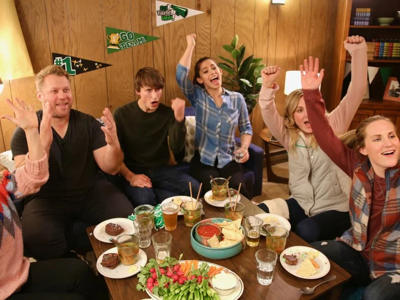 Molly Yeh, her husband Nick and their friends cheer as they watch a college hockey game on TV while eating the meal Molly prepared, as seen on Girl Meets Farm, Season 5.