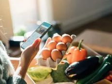 Cropped shot of woman using smartphone while picking up eggs from a delivery box filled with fresh organic fruits and vegetables at home.
