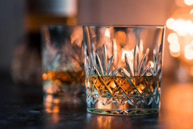 Close up color image depicting two fine crystal glasses of malt whisky on a white wooden surface. The glasses of whisky are surrounded by whisky paraphernalia such as a glass decanter and a hip flask. Room for copy space.