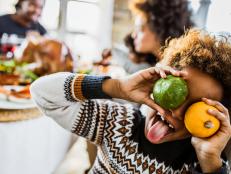 Small black girl sticking her tongue out while hiding eyes with small pumpkins at home. There are people in the background.