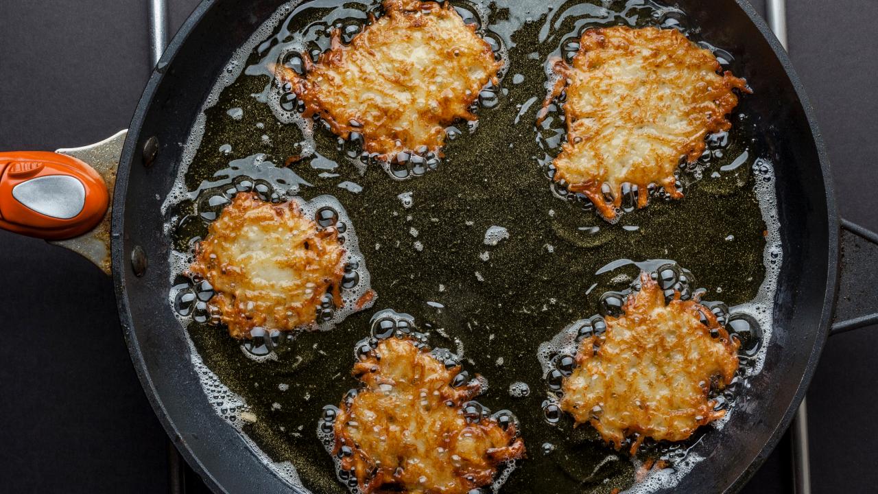 Frying latkes in a skillet, puffs of flour and oil cobwebbing out onto the  stovetop. The cook flipping the potato pancakes with a spatula, kids  eagerly peeking inside the skillet. Generative AI