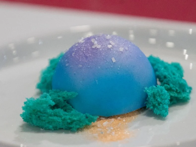 Mini Treat created by Brittani Diehl with .Purple Sugar, Blue Sugar,.Orange Sugar and Marshmallows.  It is a S'mores shaped like a Gumdrop (graham cracker crust with homemade marshmallow filling and chocolate cake, crusted in chocolate and then airbrushed, painted, and sugared to look like the Gumdrops in the land) on top an "edible moss" that looks like the ground texture in Gumdrop Land, as seen on Candyland, Season 1.